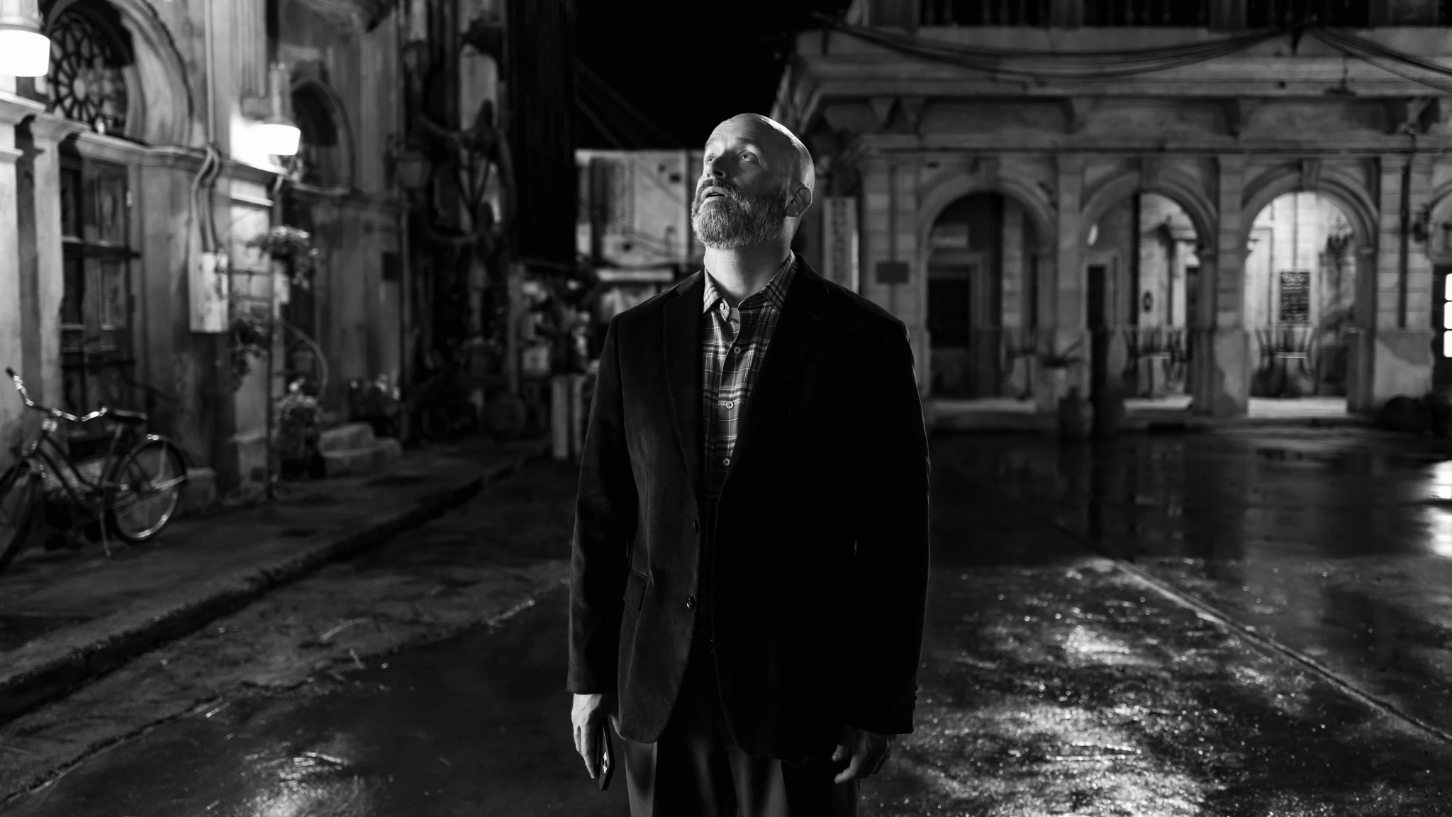 A bald, bearded man stands on a wet street in black and white, looking upward