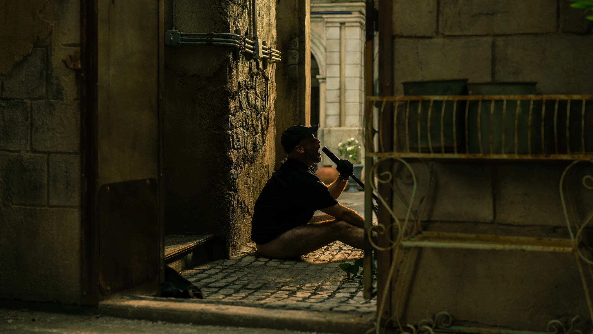 A man in a cap and black shirt sits beside a stone doorway, holding a long object