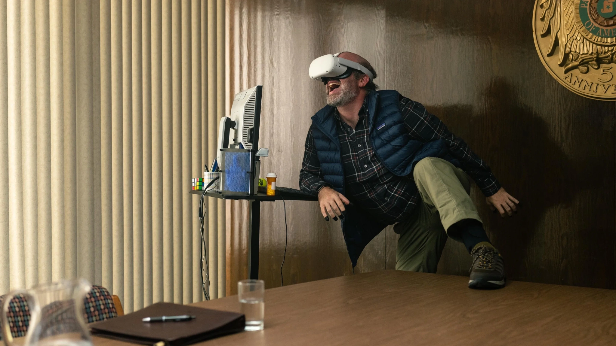A man wearing a virtual reality headset leans beside a desk and computer monitor