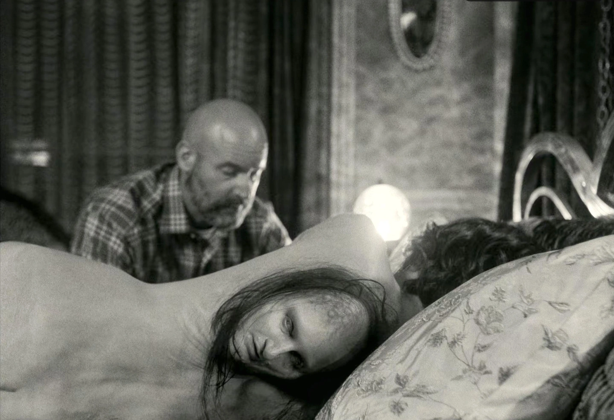 A black and white scene with two adults on a bed, one in the foreground and the other behind