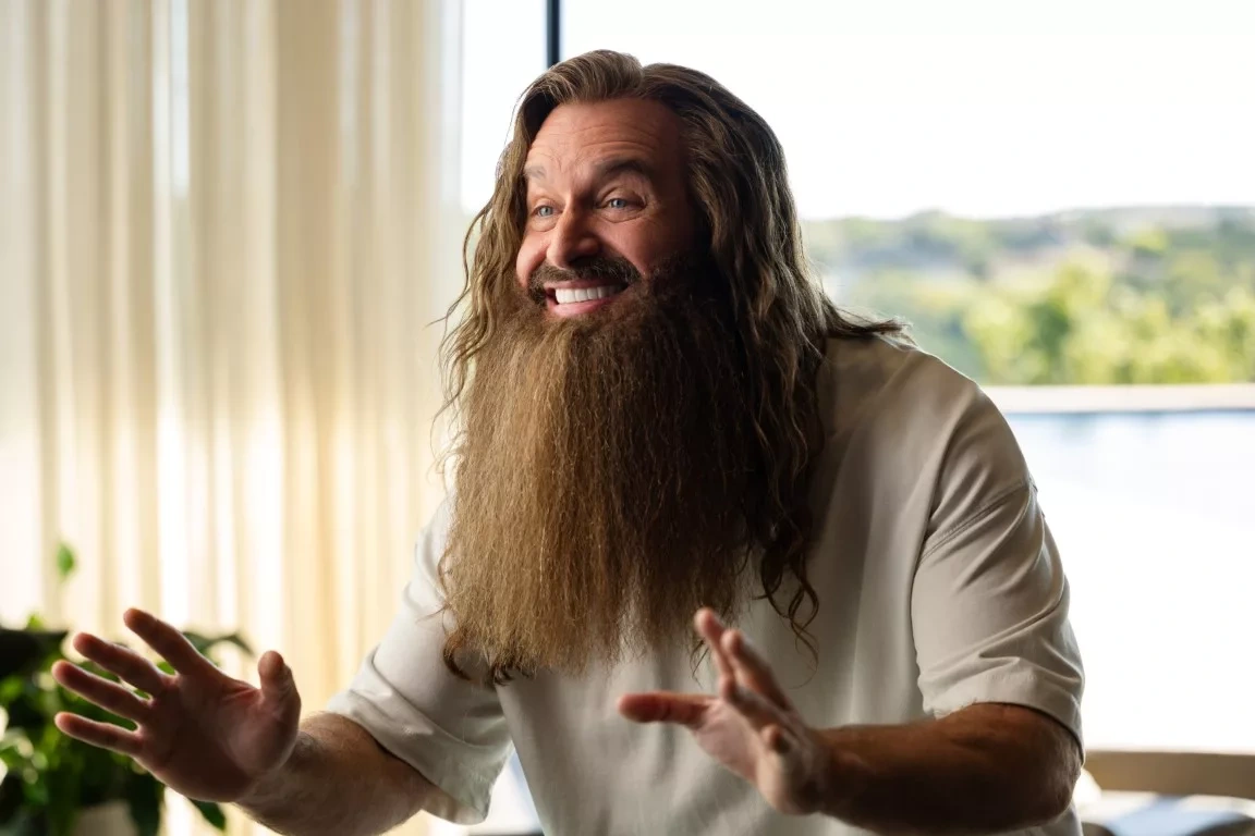A man with long hair and a full beard smiles and raises his hands in a bright room