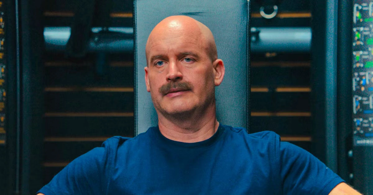 A bald man with a mustache and blue shirt sits before a background of equipment