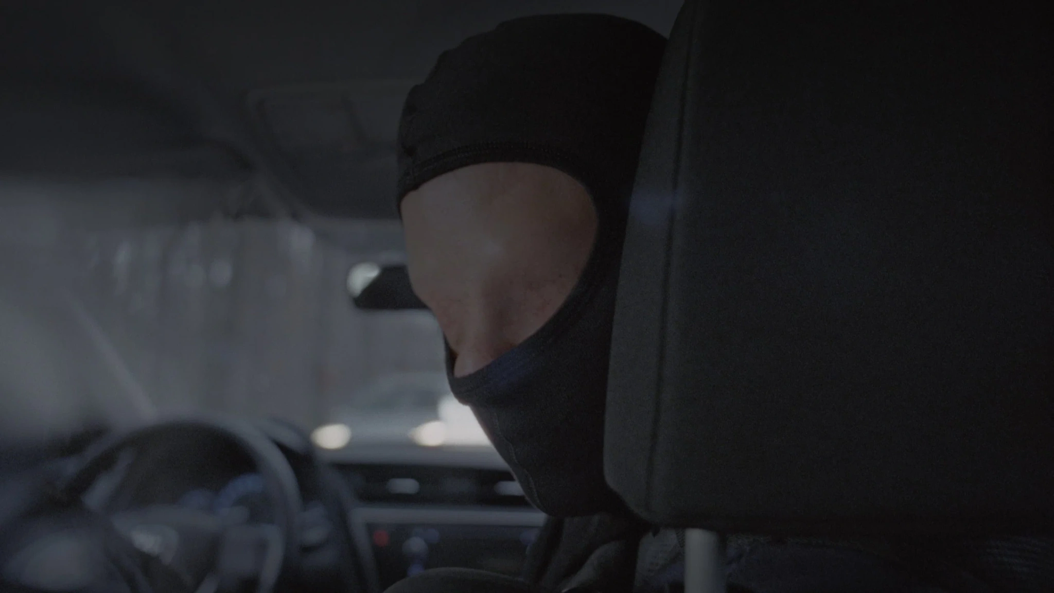 A person wearing a black hood inside a car, with the steering wheel and dashboard in the background