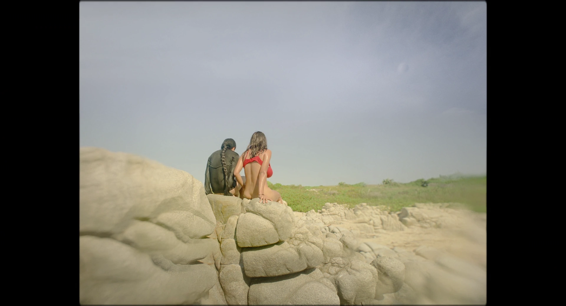 Two people sit on pale rocks beneath an overcast sky, with green land behind them