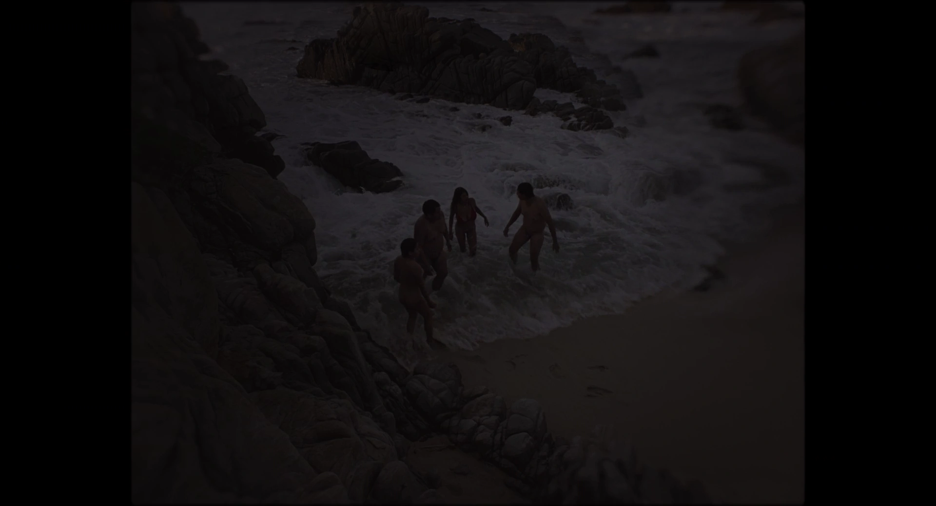 People on a rocky shore with waves breaking around them in dim light