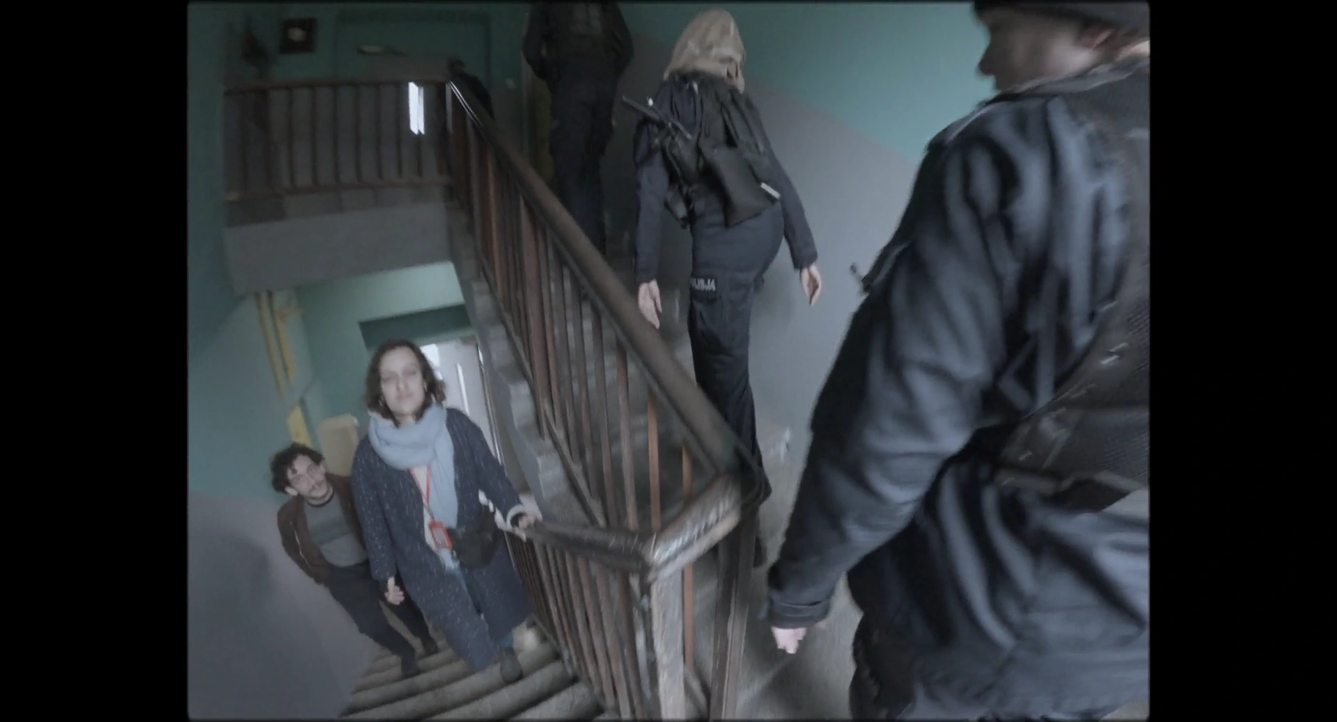 Police officers beside a staircase inside a house, with a woman looking toward them from below