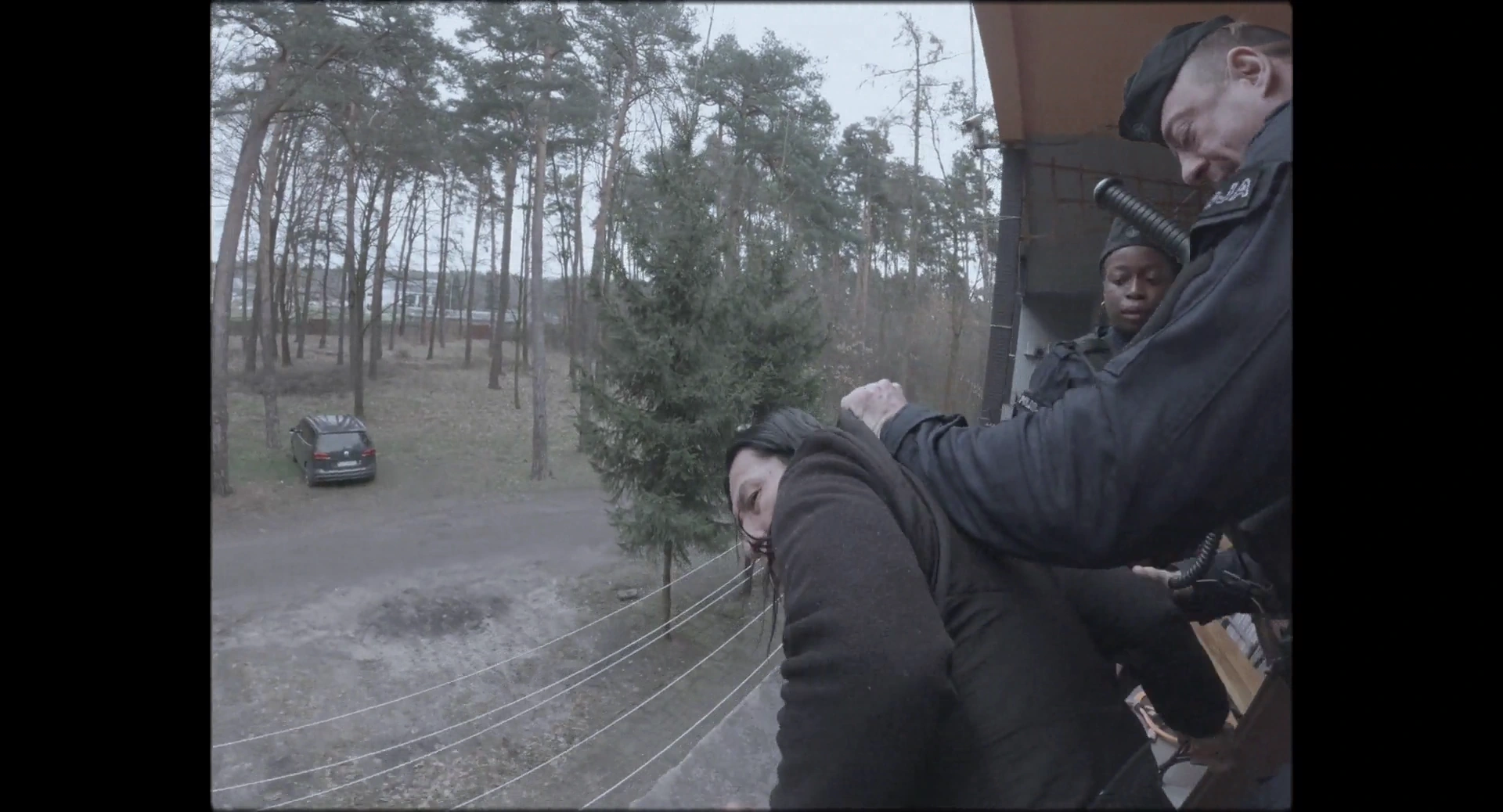 A man holds a woman behind her head near the entrance to a house surrounded by trees