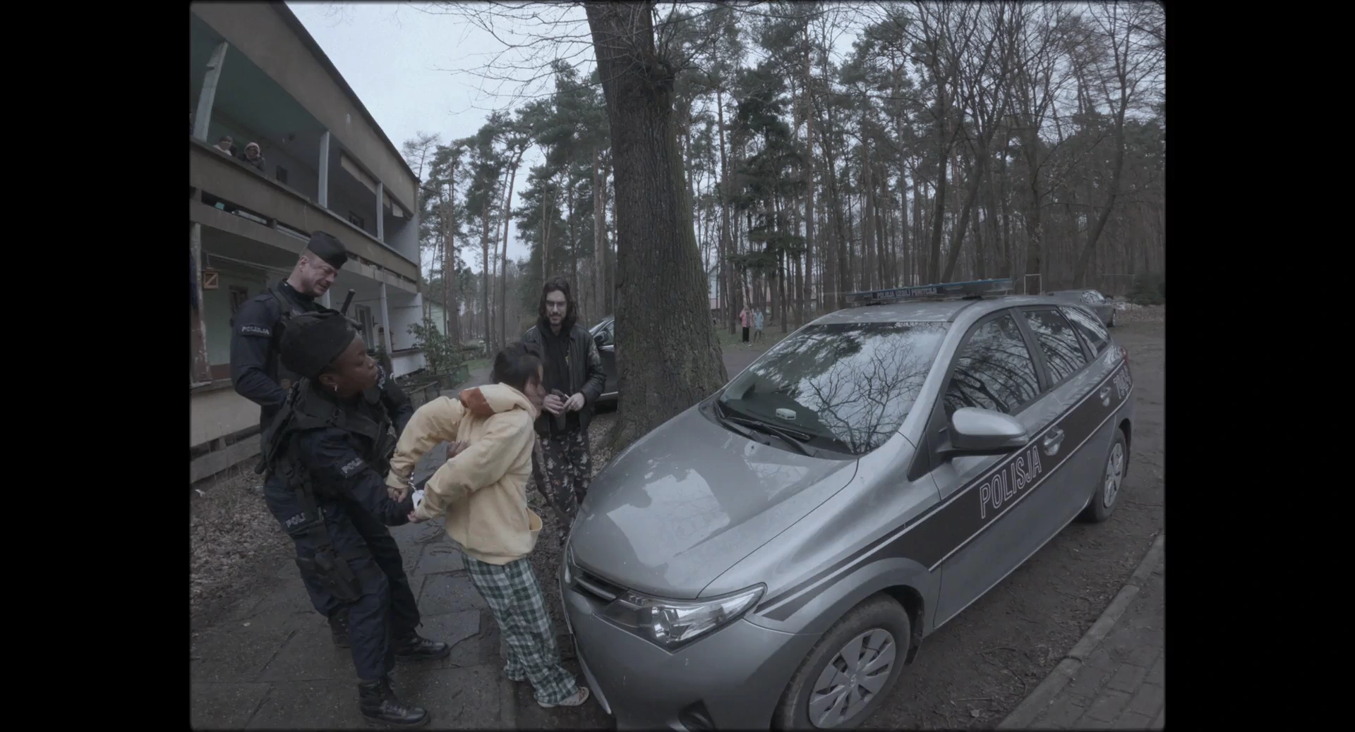 A woman in a pale covering is surrounded by men beside a parked car outside a house among trees