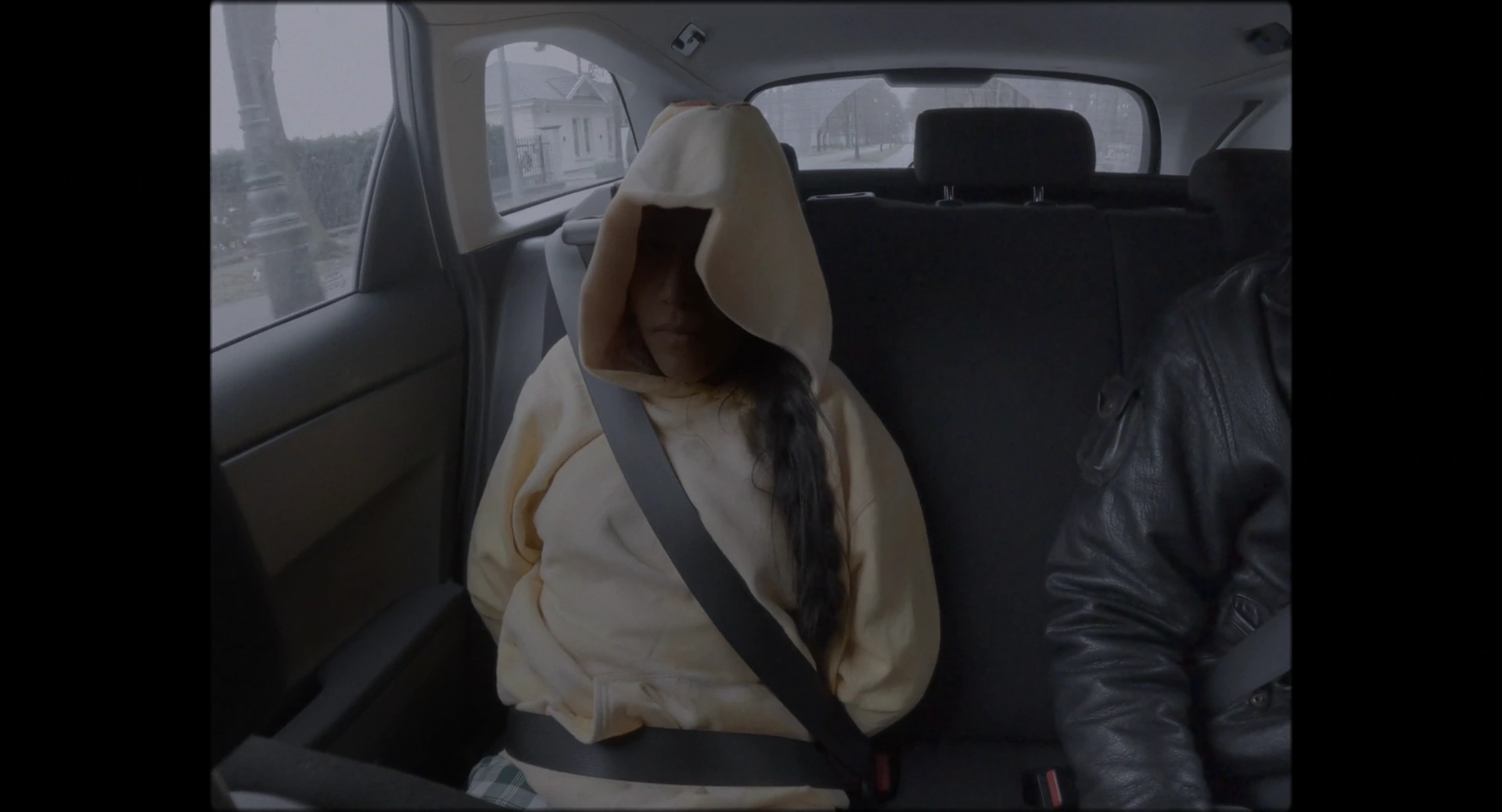 A woman sits in a car's rear seat with pale fabric covering her head and a seat belt across her