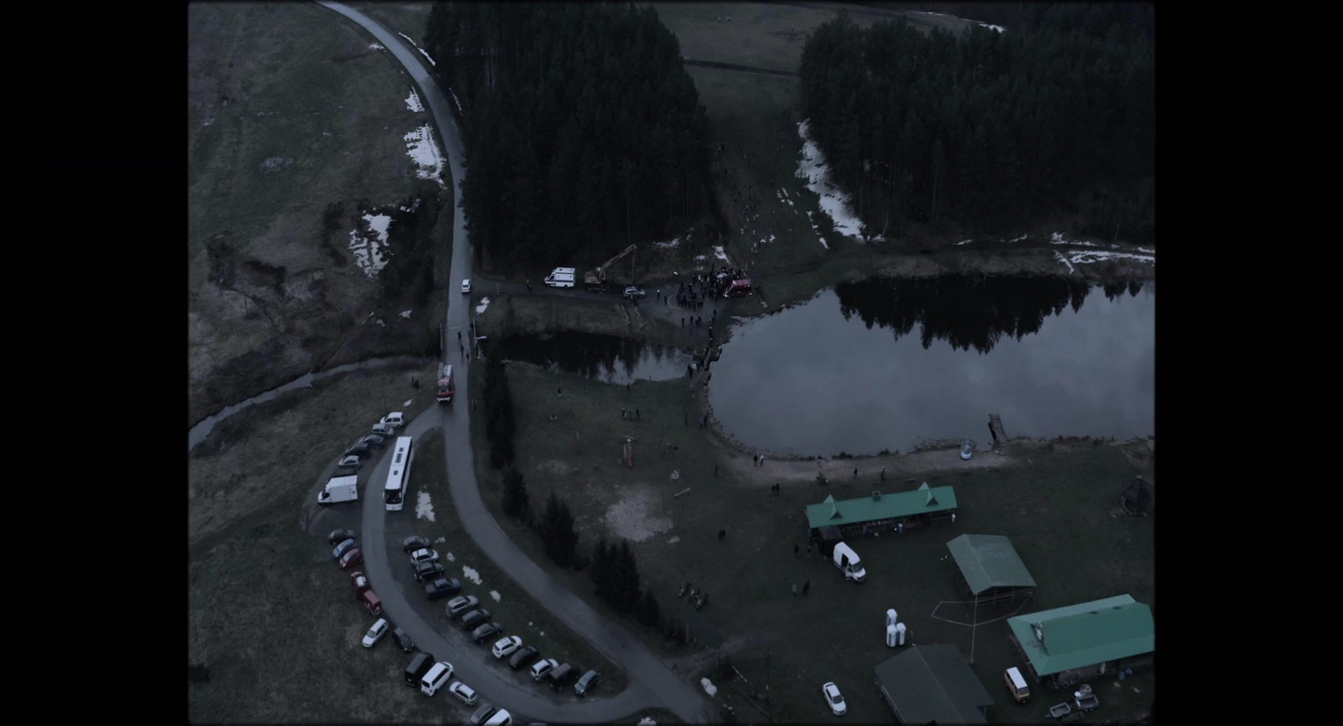 An overhead view of a lake bordered by trees, a road, cars and groups of people