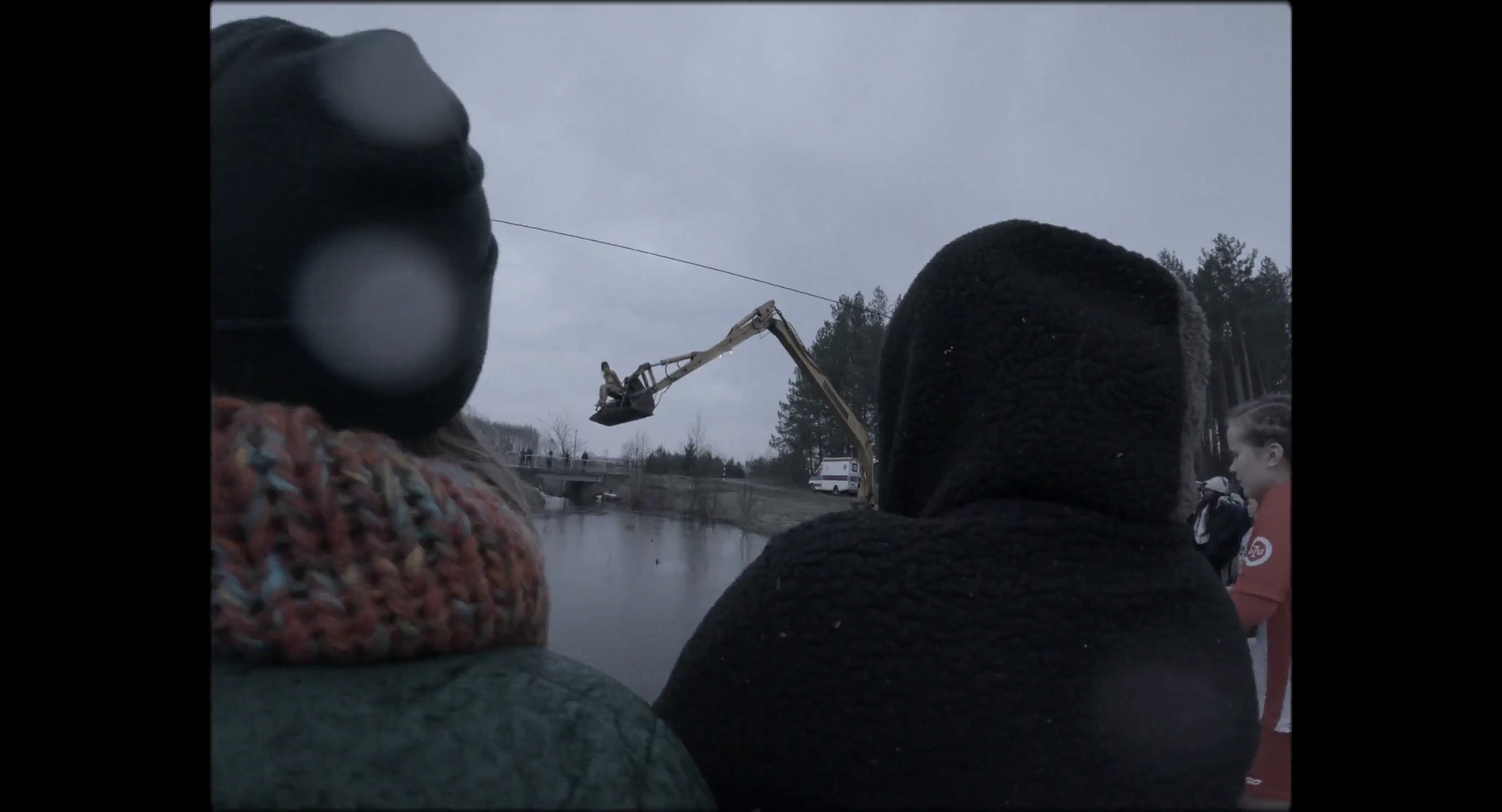 People seen from behind watch excavation equipment extending over water beneath an overcast sky
