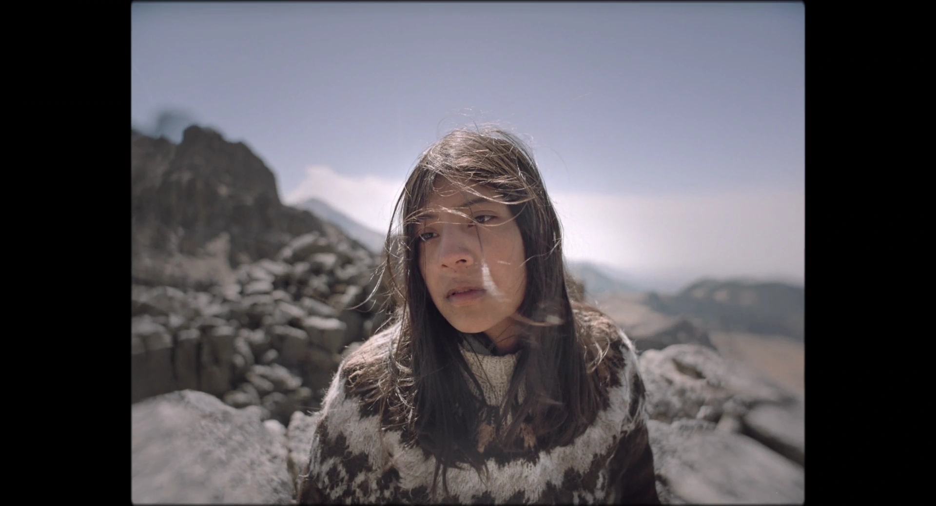 A dark haired woman before rocks and mountains looks beside the camera in daylight