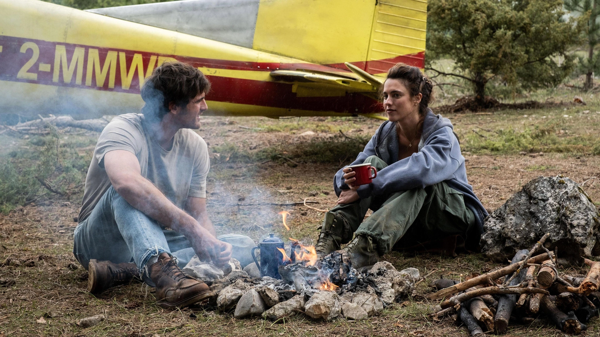 Hig and Cima sit beside a small fire and cooking utensils, with the yellow plane behind them
