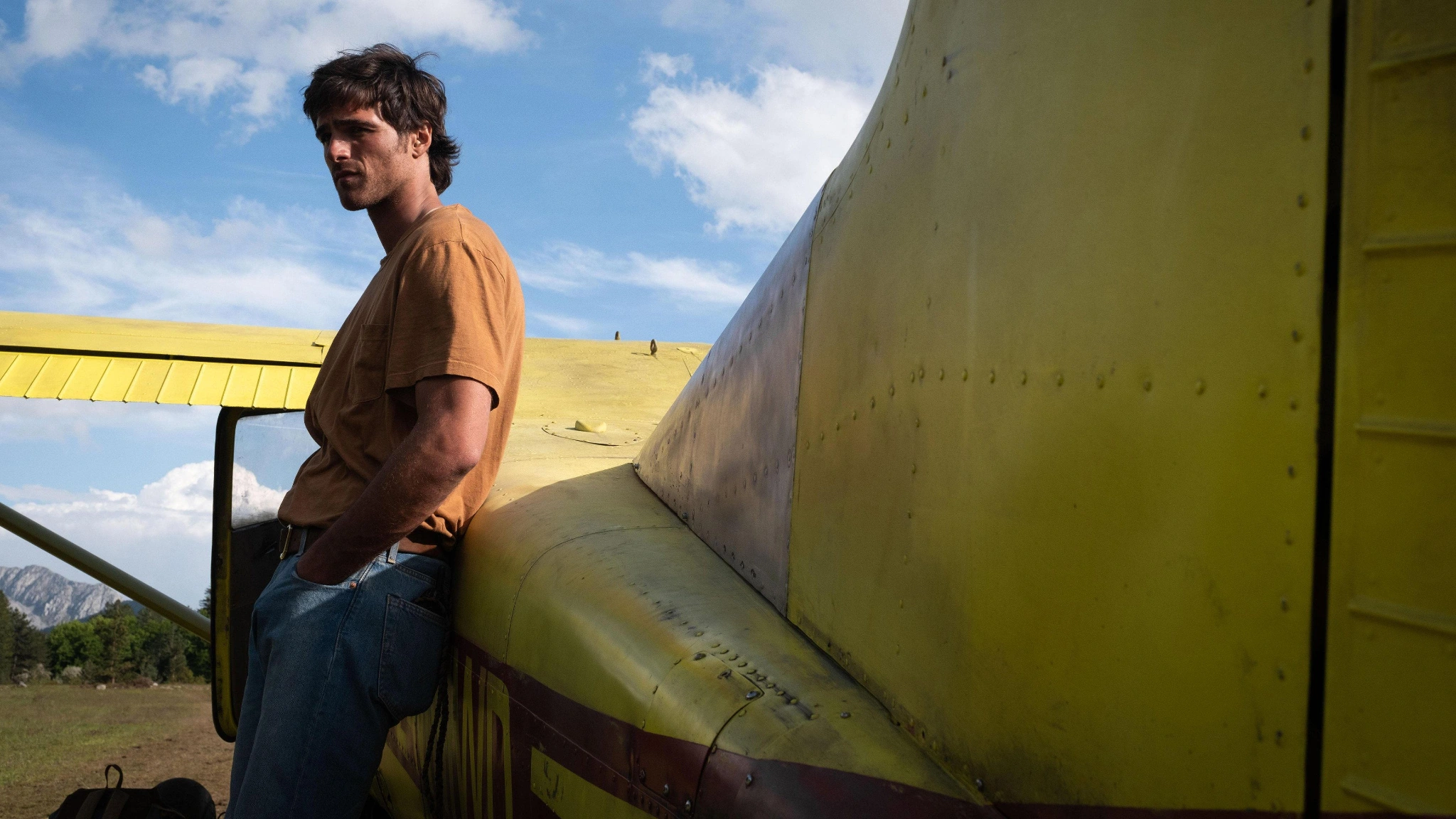 Hig leans against the body of a yellow plane beneath a blue sky near mountains