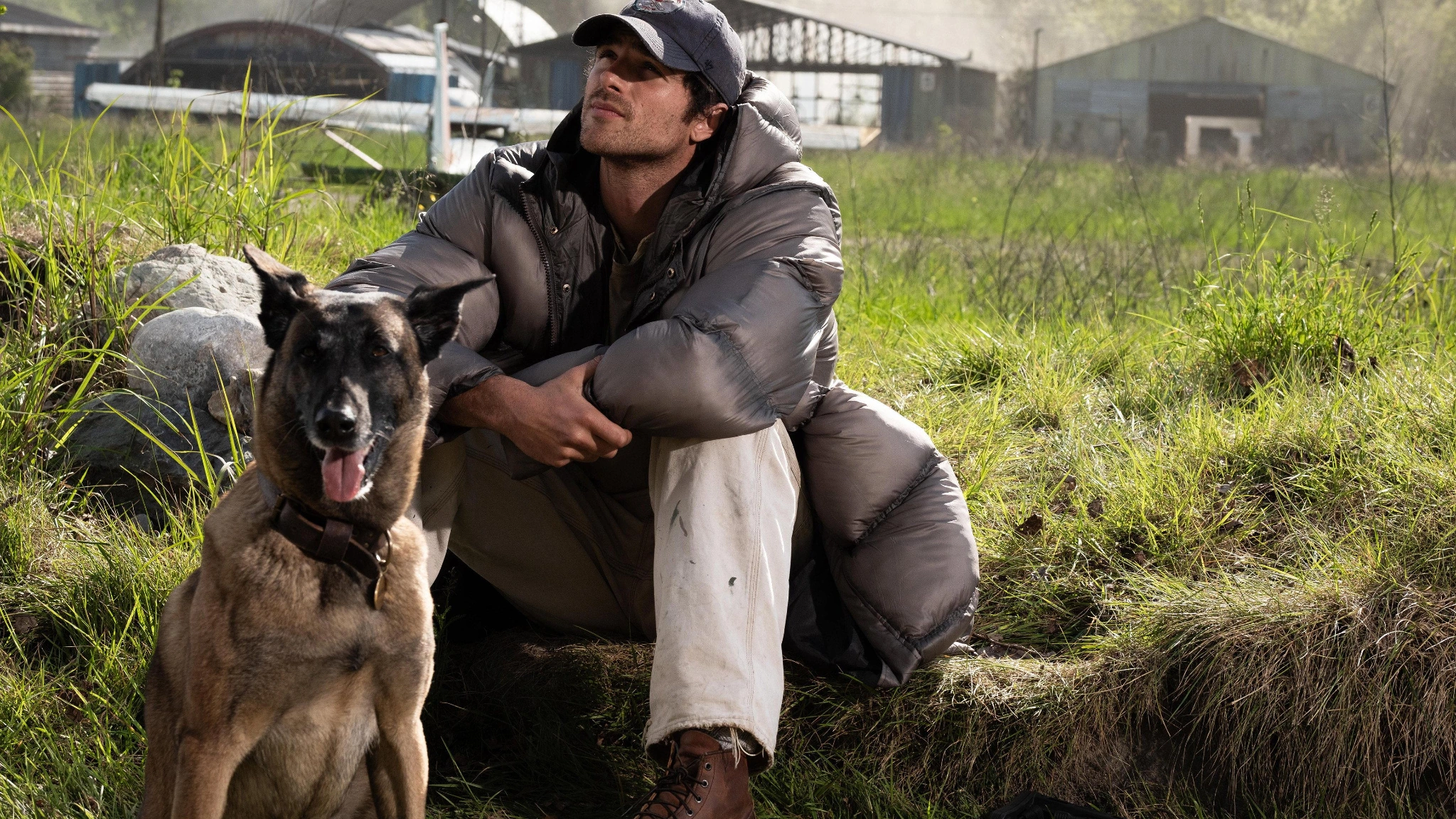 Hig sits beside Jasper on grassy ground before buildings and hangars near the airfield