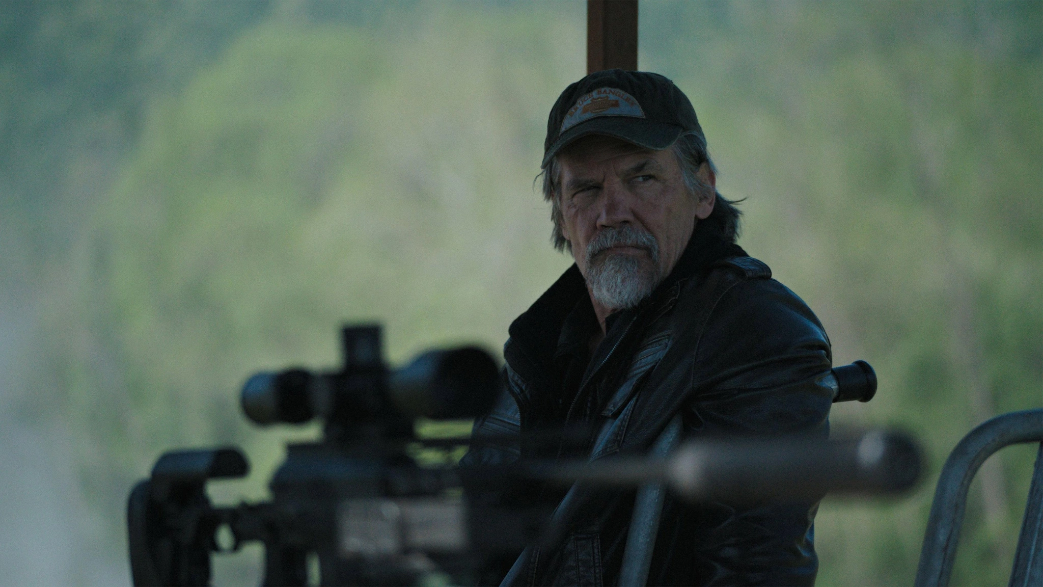 Bangley wears a cap and dark jacket while sitting behind a scoped rifle