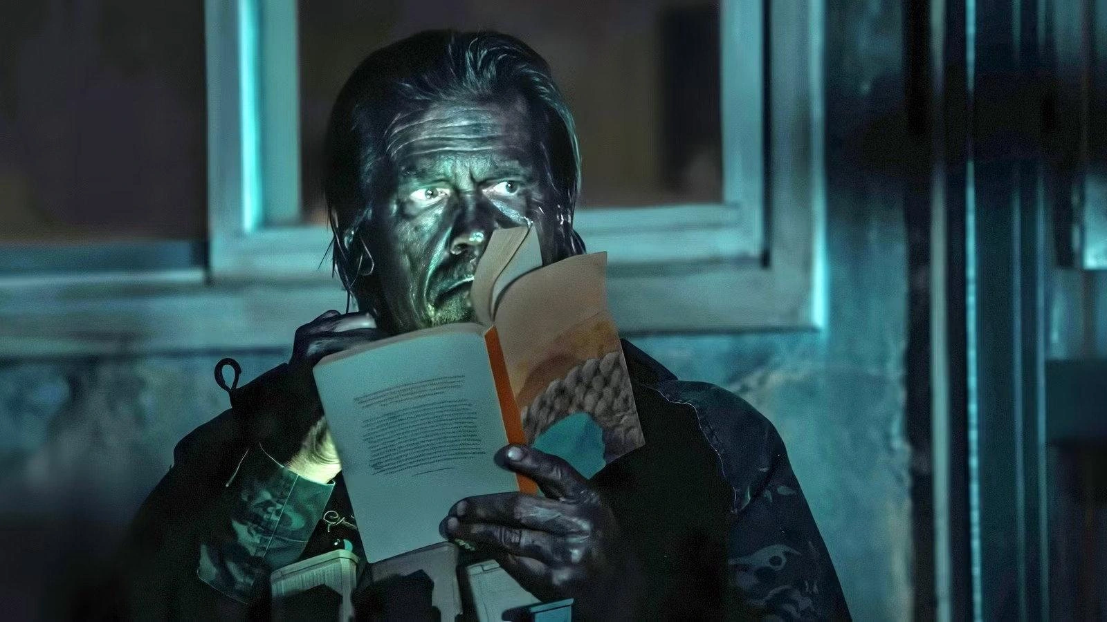 A man holds an open book near his face and looks upwards under cold blue lighting