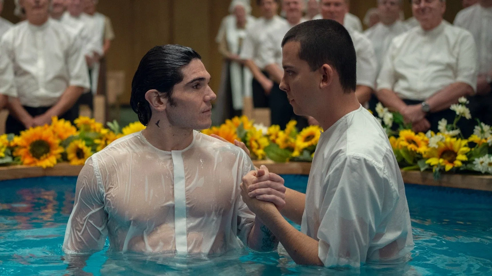 Two men in white shirts stand in a pool before other men and yellow flowers