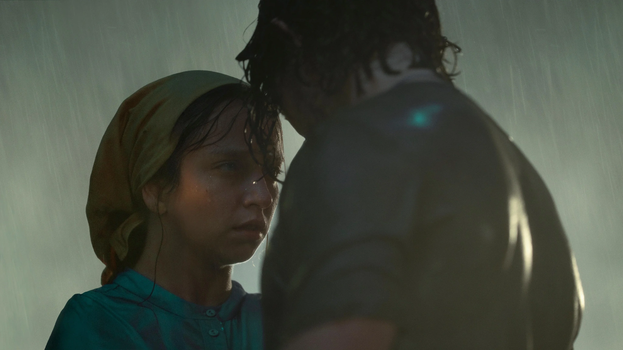 A woman in a head covering and blue shirt looks toward a man standing close to her in the rain