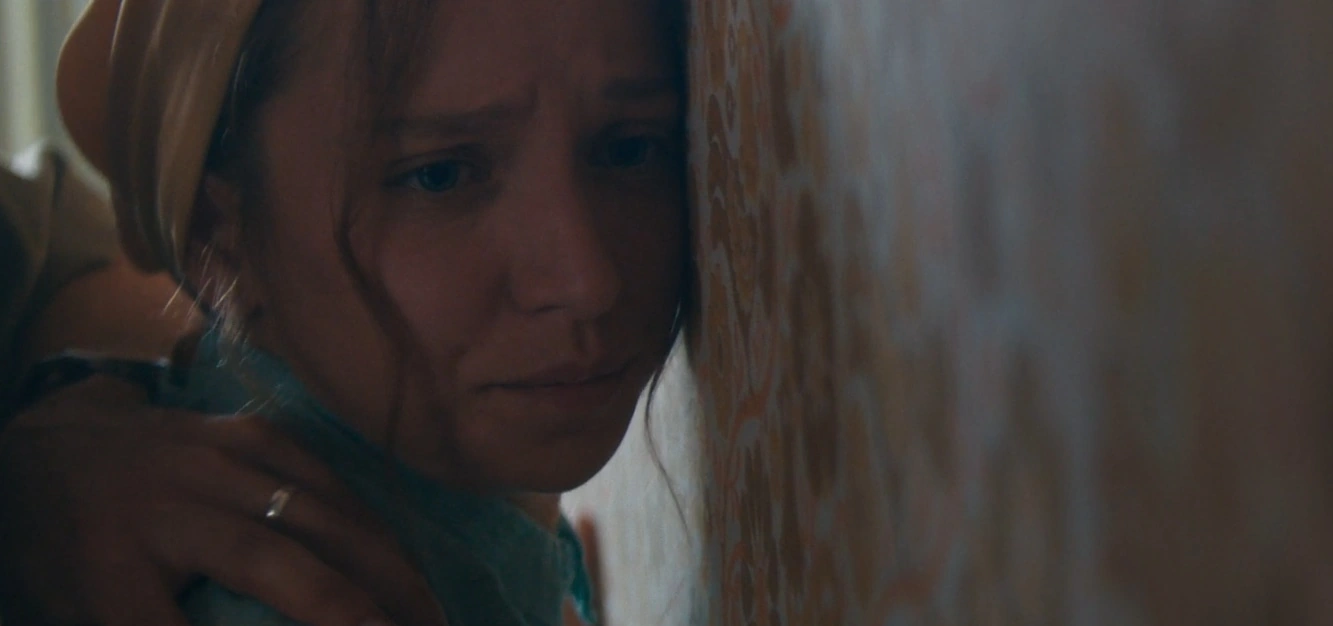 A tearful woman beside a patterned curtain with a hand resting on her shoulder