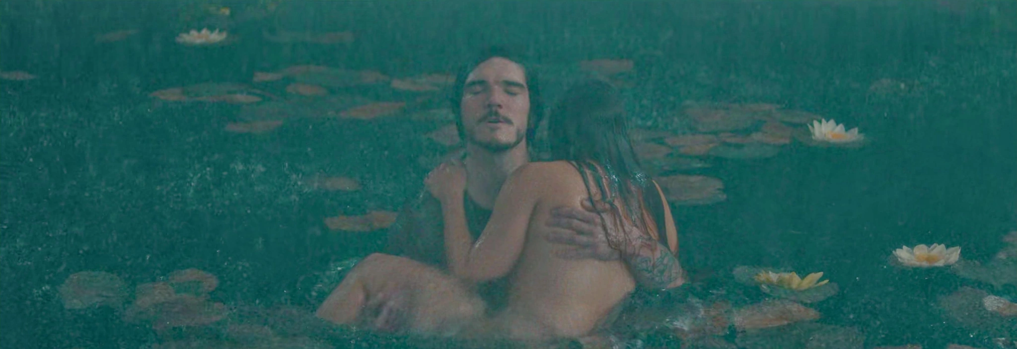 Two adults close together in water, one seen from behind, surrounded by leaves and water flowers