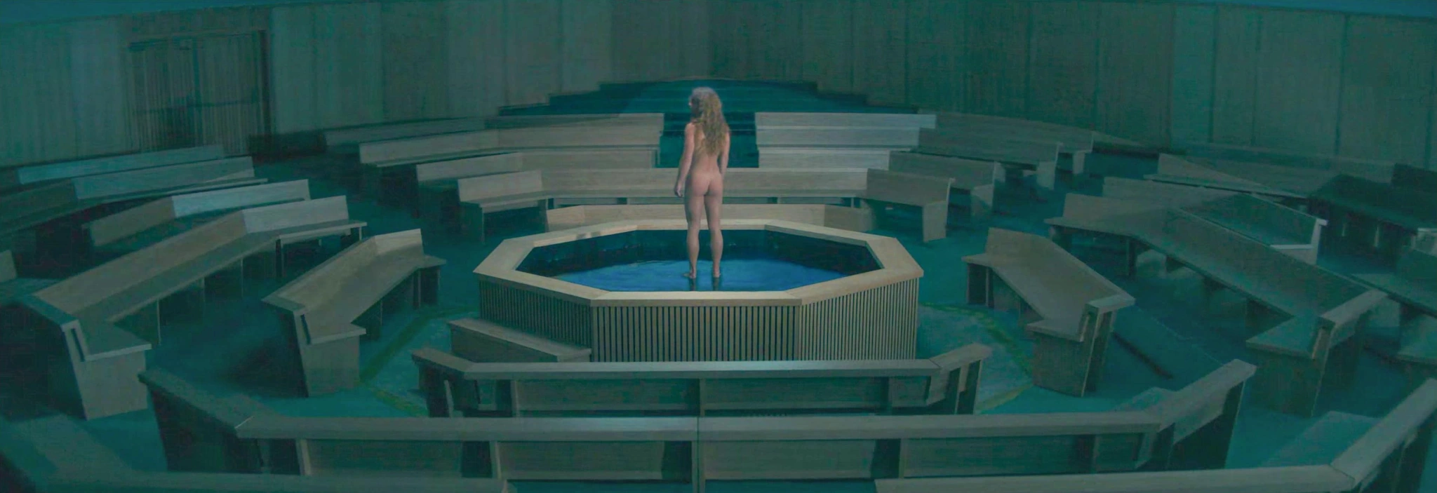 A nude adult woman seen from behind stands in a pool at the center of a hall with circular wooden seating